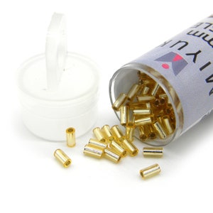 Puede incluir: Un pequeño tubo de plástico transparente lleno de cuentas de semillas doradas. El tubo tiene una etiqueta blanca con el texto "MIYU 3mm Bugle".