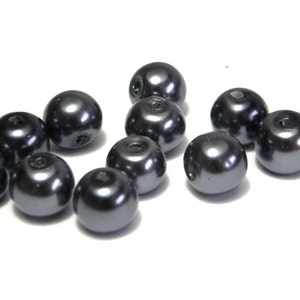 May include: A collection of dark gray, round beads with a glossy finish. Each bead has a small hole visible at the top. The beads are scattered on a white surface, creating a visually appealing arrangement. These beads are suitable for jewelry making.