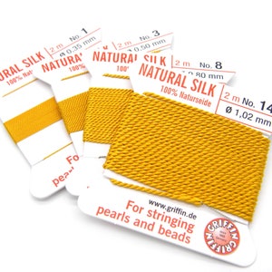 May include: Four spools of yellow natural silk thread for stringing pearls and beads. The spools are labeled with the brand name "Griffin" and the thread thickness in millimeters. The text on the spools reads "Natural Silk 100% Naturseide" and "For stringing pearls and beads".