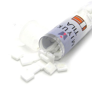 May include: A transparent plastic tube, partially open, filled with small, white, square beads. Some beads are scattered around the tube. The tube is labelled with the text "MIYUKI TILA". These beads are likely for crafts or jewellery.