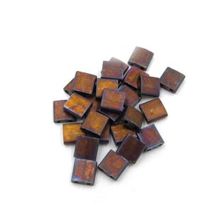 Pode incluir: Um conjunto de contas quadradas com acabamento metálico. As contas apresentam uma mistura de tons castanhos, dourados e roxos. Estão espalhadas sobre um fundo branco, mostrando as suas formas e variações de cor.