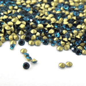 May include: Close-up of a collection of teal and gold rhinestones. The teal stones are round and faceted, while the gold stones have a faceted, pyramid shape. The rhinestones are scattered across a white surface.