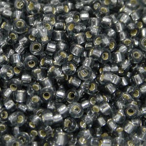 Pode incluir: Close-up de uma pilha de pequenas contas cilíndricas. As contas são de cor cinza translúcido com um orifício de cor dourada metálica que atravessa o centro. As contas estão espalhadas, preenchendo a moldura.