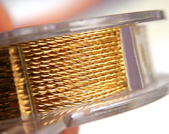 May include: Close-up of a spool of gold-colored wire. The wire is tightly wound on a clear plastic spool. The wire has a textured, braided appearance, and reflects light, creating a shiny effect. The spool is partially visible.