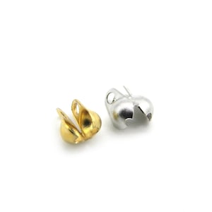 Pode incluir: Duas tampas de contas de metal, uma dourada e outra prateada, são exibidas em fundo branco. As tampas têm formato arredondado com design dividido e um pequeno laço para fixação.