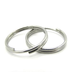 Pode incluir: Dois chaveiros divididos prateados, sobrepostos sobre um fundo branco. Cada anel é feito de uma faixa de metal arredondada de camada dupla, com uma pequena abertura para adicionar chaves ou outros itens. Os anéis são simples e funcionais.