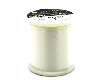 Può includere: Una bobina di filo per perline MIYUKI bianco sporco, etichettata "NI 1 OR". La bobina è cilindrica con base e parte superiore in plastica bianca, e l'etichetta è in bianco e nero. Il filo è ben avvolto.