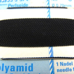 Può includere: Primo piano di un filo nero avvolto attorno a una carta bianca con testo blu. Il testo recita "Polyamid 1 Nadel needle fixed".