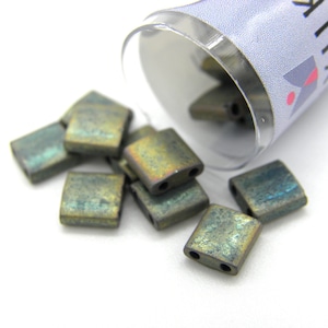 Può includere: Un piccolo contenitore di perline quadrate di vetro verde e marrone iridescente. Il contenitore è in plastica trasparente e ha un'etichetta bianca con la parola "Ruby" stampata su di essa.