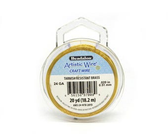 May include: A spool of gold-colored Beadalon Artistic Wire in tarnish-resistant brass. The label indicates 24 gauge, .020 in diameter, and a length of 20 yd. The clear plastic packaging features a hanging tab and a barcode for identification.