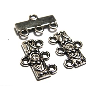 May include: Close-up of three silver-coloured metal jewellery connectors. Two connectors feature floral and circular designs, while the third is a simple rectangular bar. All have multiple loops for attaching to other components.
