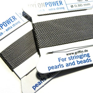 Könnte beinhalten: Zwei Spulen grauer Nylon-Schnur zum Fädeln von Perlen und Perlen. Die Spulen sind mit "Nylon Power" und "Kein Stretch - extra stark" beschriftet. Die Schnur ist 0,80 mm dick.
