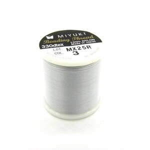 Pode incluir: Um carretel de fio de miçangas Miyuki cinza claro. O rótulo diz "Beading Thread" e "100% Nylon Made in Japan". O carretel é rotulado como "330dtex" e "MX25R".