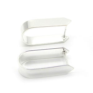 Pode incluir: Um par de brincos de argola prateados. Cada brinco tem uma parte superior retangular e uma base curva, criando um design aberto único. Os brincos são apresentados em um fundo branco.