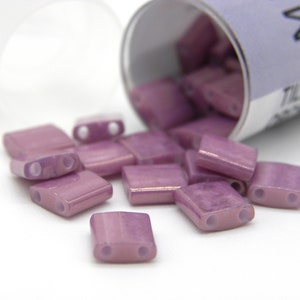 Può includere: Un contenitore di piccole perline di vetro rettangolari di colore lilla chiaro con due fori. Le perline hanno una lucentezza metallica.