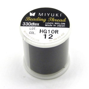 Könnte beinhalten: Eine Spule schwarzes Miyuki-Perlenfaden. Das Etikett lautet "Beading Thread" und "100% Nylon Made in Japan". Die Spule trägt auch die Aufschrift "330dtex", "LOT. HG1OR 12" und "MIYUKI CO. LTD."