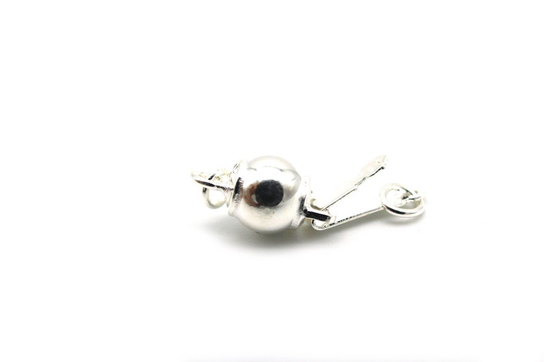 Może przedstawiać: Srebrne zapięcie do biżuterii, prawdopodobnie do naszyjnika lub bransoletki, na białym tle. Zapięcie posiada okrągły, kulisty element i zawiasowe zamknięcie dźwigniowe. Metal ma jasne, odblaskowe wykończenie.