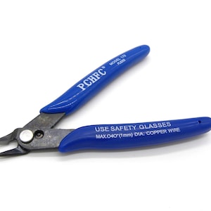 Può includere: Tronchesi con manici blu con la scritta "PCHFC" e "MODEL 170 PLIERS". L'utensile ha un tagliente in metallo argentato. È visibile anche il testo "USE SAFETY GLASSES MAX.040"(1mm) DIA. COPPER WIRE".