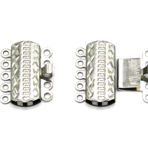 Puede incluir: Dos cierres de metal plateados, cada uno con cuatro bucles, diseñados para sujetar cadenas o cordones de joyería. Los cierres presentan un patrón decorativo y un cierre de resorte.