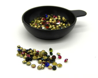 May include: A black, handled bowl filled with a variety of colorful rhinestones. The rhinestones are in shades of gold, red, blue, green, and black. Some rhinestones are scattered on the white surface.