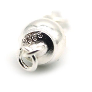 Może przedstawiać: Zbliżenie małego, srebrnego charmsa w kształcie dzwonka. Charms ma pętlę do mocowania, a na powierzchni wytłoczony jest napis "S925". Tło jest białe.