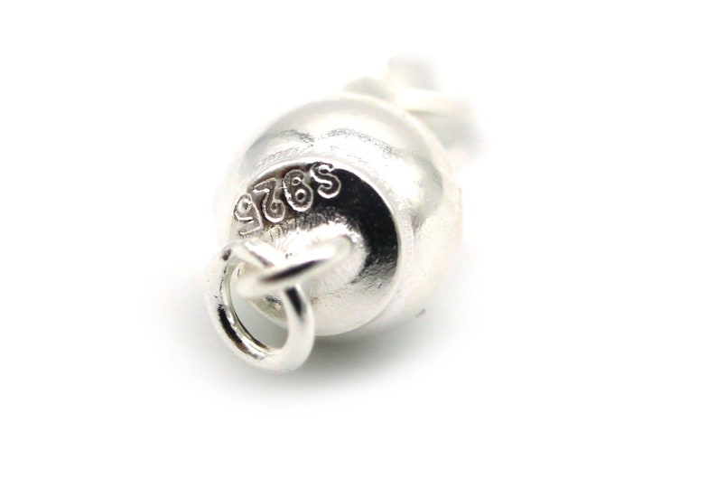 Może przedstawiać: Zbliżenie małego, srebrnego charmsa w kształcie dzwonka. Charms ma pętlę do mocowania, a na powierzchni wytłoczony jest napis "S925". Tło jest białe.