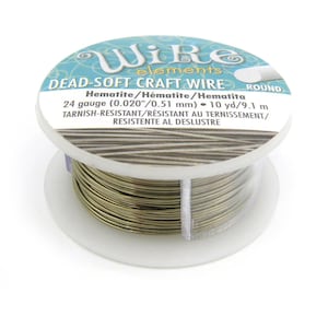 Puede incluir: Una bobina de alambre redondo para manualidades Wire Elements de color hematita. La etiqueta indica calibre 24, 0,51 mm y 9,1 m de alambre resistente al deslustre. La bobina es de plástico blanco con una etiqueta azul y blanca.