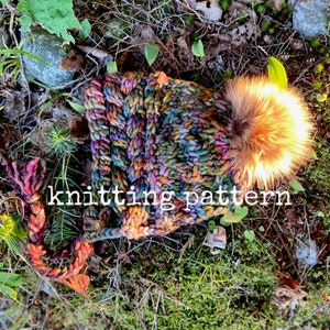 Op de afbeelding: Een kleurrijke gebreide muts met een pluche pompon. De muts is gemaakt van een dik garen en heeft een gevlochten band. De muts ligt op een bed van mos en bladeren.