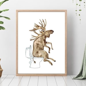 Puede incluir: Impresión en acuarela de un alce sentado en un inodoro. El alce es marrón con grandes astas. El inodoro es blanco y la obra de arte está enmarcada en un marco de madera clara. La obra de arte se exhibe en una habitación con una planta y una silla.