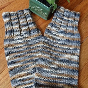 Puede incluir: Un par de calentadores de brazo de punto a rayas en tonos grises, marrones y blancos. Los calentadores de brazo están doblados y colocados sobre una superficie de madera. El texto "*** KNIT PATTERN ***" es visible en la parte inferior de la imagen.