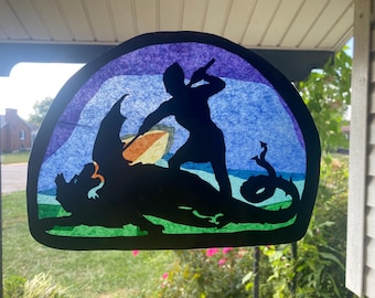 May include: A stained glass window decoration depicting a knight slaying a dragon. The knight is silhouetted in black and is holding a sword. The dragon is also silhouetted in black and has a green and blue background.