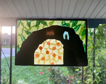 May include: A stained glass window depicting a cave with a green leafy background. The cave is black with a yellow and orange patterned opening.