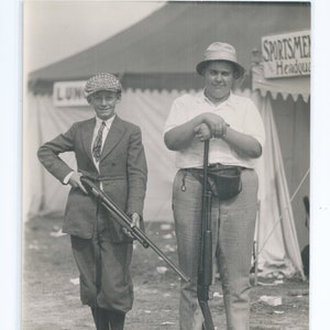 May include: Black and white photograph of two people, each holding a long gun. One person wears a tweed cap, suit, and tie. The other wears a hat, white shirt, and pants. A tent is in the background with the words "SPORTSMEN Headquarters".