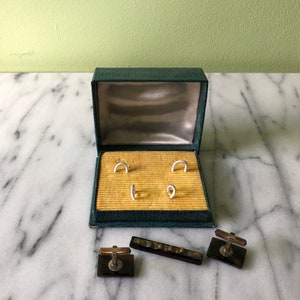 May include: An open, vintage green jewelry box with a gold interior holds four silver-toned cufflink studs. Two matching cufflinks and a tie bar with dark brown and gold accents are displayed outside the box on a marble surface.