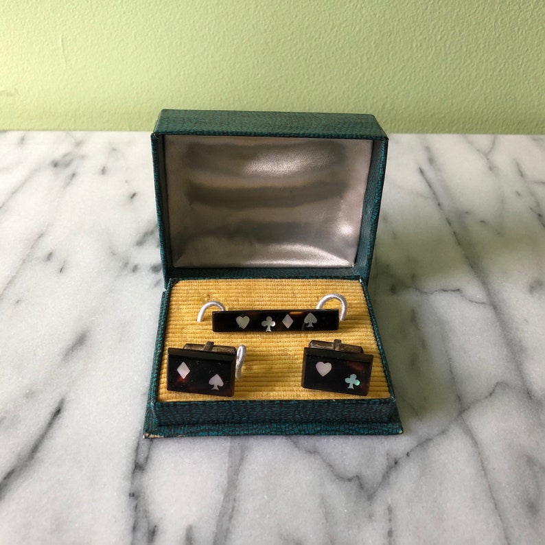 May include: A vintage set of playing card-themed accessories, including cufflinks and a tie bar, presented in a green box. The accessories feature playing card suit symbols: hearts, diamonds, clubs, and spades. The box has a gold interior.