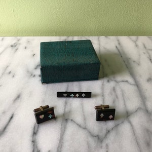 May include: A vintage set of playing card suit-themed accessories, including cufflinks, a tie bar, and a green box. The cufflinks and tie bar are black with inlaid heart, diamond, club, and spade symbols.