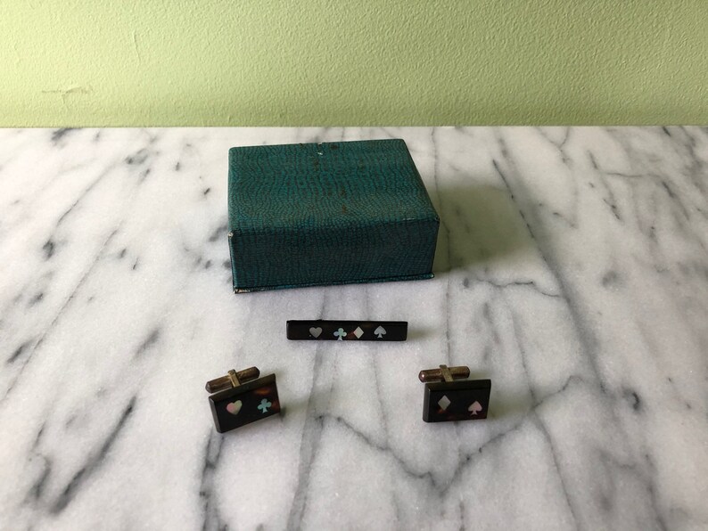 May include: A vintage set of playing card suit-themed accessories, including cufflinks, a tie bar, and a green box. The cufflinks and tie bar are black with inlaid heart, diamond, club, and spade symbols.