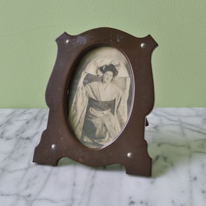 May include: A dark brown wooden picture frame with an oval opening. The frame has a decorative design and is standing on a white marble surface. A black and white photograph of a woman in a dress is inside the frame.
