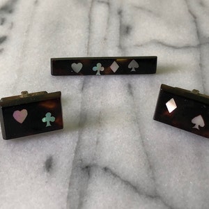May include: A set of vintage playing card-themed accessories, including a tie bar and cufflinks. The pieces feature a tortoiseshell pattern with inlaid mother-of-pearl suit symbols: hearts, clubs, diamonds, and spades. The cufflinks are rectangular.