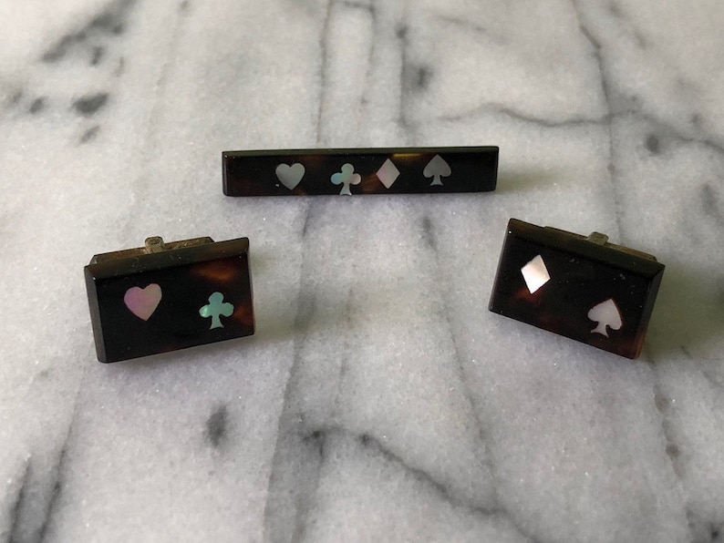 May include: A set of vintage playing card-themed accessories, including a tie bar and cufflinks. The pieces feature a tortoiseshell pattern with inlaid mother-of-pearl suit symbols: hearts, clubs, diamonds, and spades. The cufflinks are rectangular.