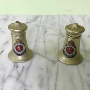 May include: A pair of vintage silver-toned salt and pepper shakers. Each shaker has a flared base and a small, round top. A colorful emblem with text is affixed to the front of each shaker. The shakers are displayed on a marble surface.