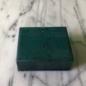 May include: A square, teal-colored box with a textured, reptile-skin pattern. The box is closed and appears to be made of cardboard or a similar material. The box is sitting on a white and gray marble surface.