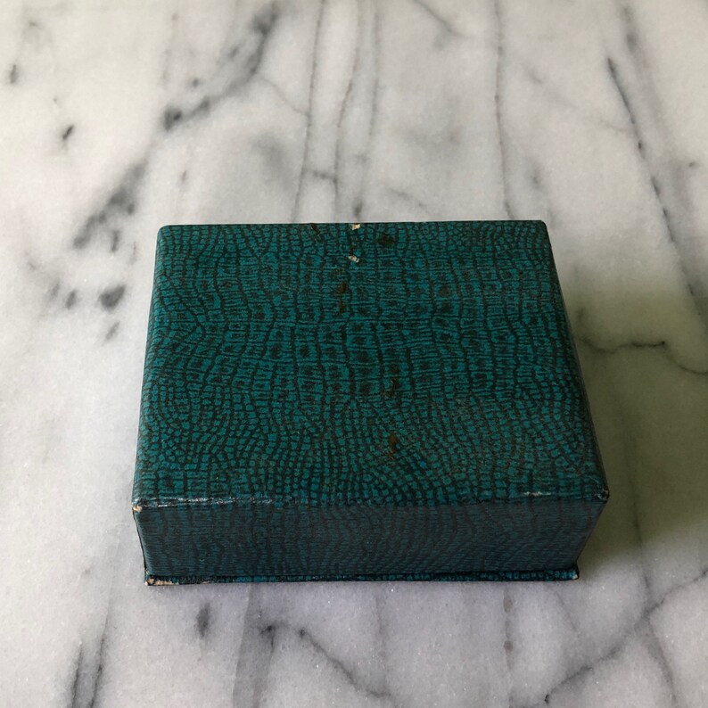 May include: A square, teal-colored box with a textured, reptile-skin pattern. The box is closed and appears to be made of cardboard or a similar material. The box is sitting on a white and gray marble surface.
