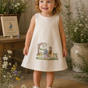 May include: A sleeveless cream-colored dress with an embroidered scene from "The Tale of Peter Rabbit" is worn by a young child. The child is wearing white shoes and a white hair bow. A copy of the book is visible in the background.