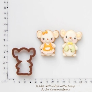 Peut inclure: Deux emportes-pièces en forme de souris, l'un avec une robe jaune et l'autre avec un gilet vert. Les emportes-pièces sont à côté de deux biscuits décorés, l'un avec une robe jaune et l'autre avec un gilet vert. Les biscuits sont sur une surface blanche avec une règle en centimètres.