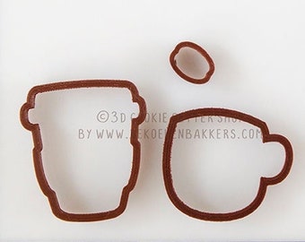 May include: Brown cookie cutters in the shape of a coffee cup, a to-go coffee cup, and a coffee bean. The cutters are on a white surface with a ruler in centimeters. The text "3D COOKIE CUTTER SHOP BY WWW.DEKOEKENBAKKERS.COM" is visible.