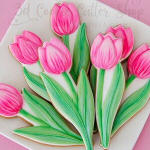May include: A bouquet of pink and green sugar cookies decorated to look like tulips. The cookies are arranged on a white plate with a pink background. The text "3D Cookie Cutter Shop" is visible in the top left corner of the image.