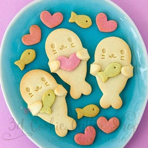 Könnte beinhalten: Ein blauer Teller mit drei Robben-förmigen Keksen, die ein Herz und einen Fisch halten. Die Kekse sind von kleineren Herz- und Fisch-förmigen Keksen umgeben.