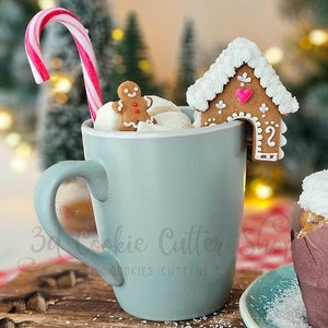 Pode incluir: Uma caneca azul com uma casa de gengibre, um homem de gengibre e um bastão de caramelo saindo dela. A caneca está sobre uma superfície de madeira com granulado branco. O texto "3D Cookie Cutter Shop" é visível na caneca.