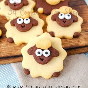 Puede incluir: Primer plano de cinco galletas de azúcar decoradas con forma de oveja. Las galletas son marrones y amarillas con glaseado blanco y ojos comestibles negros. Las galletas están sobre una tabla de cortar de madera. El texto "3d Cookie Cutter Shop" es visible en la imagen.